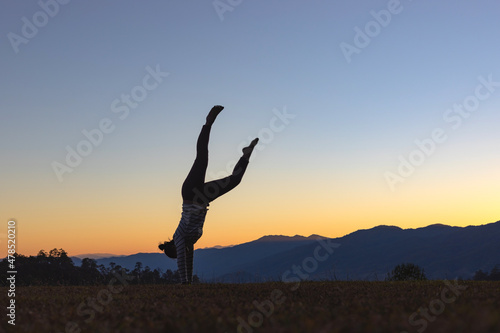 Silhouette of female gymnast playing upside down outdoors in summer park walking on hands at sunset