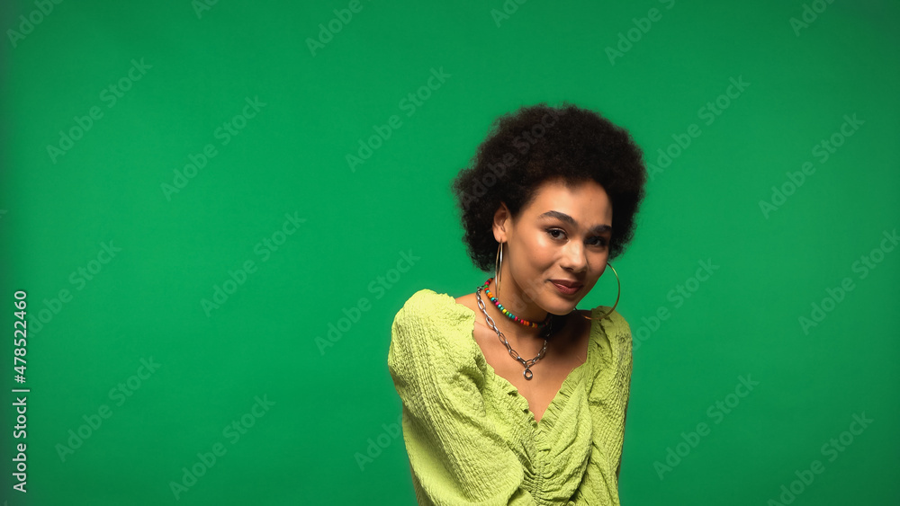 Fototapeta premium positive african american woman in blouse and hoop earrings smiling isolated on green