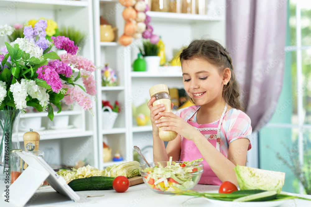 Cute little girl preparing fresh salad on kitchen table with tablet Cute little girl preparing fresh salad on kitchen table with tablet