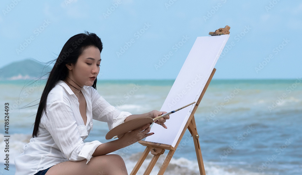 Young woman wearing white shirt standing on the beach and painting,