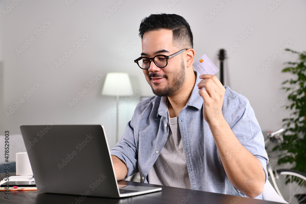 Feeling happy and confident. Young Asian man using laptop computer holding credit card for online shopping and payment at home. Feeling happy and confident. Young Asian man using laptop computer holding credit card for online shopping and payment at home.
