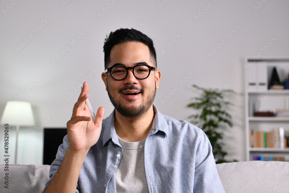 Portrait shot of happy friendly and confident Asian businessman sitting on a couch at home, looking and talking at camera and waving hand saying hello during video call Portrait shot of happy friendly and confident Asian businessman sitting on a couch at home, looking and talking at camera and waving hand saying hello during video call