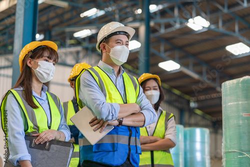 Group of Asian male and female engineers team wearing hygienic mask protect with helmet safety stand in line in factory warehouse
