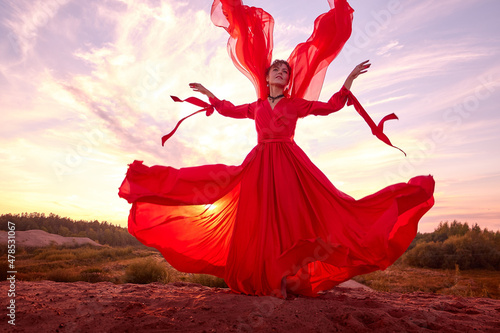 Beautiful young woman or girl with curly hair and in red dress with a light flying fabric on the sand on sunny day with blue sky in the background. Model or dancer posing in photo shoot on sand dunes