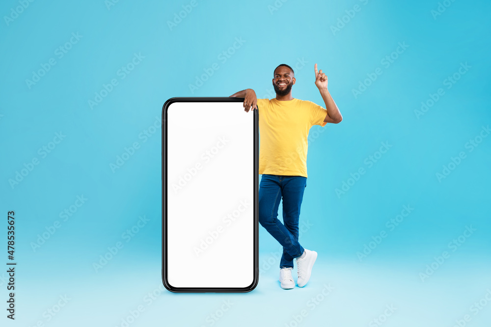 Handsome black guy offering mockup for new mobile app on giant smartphone screen, pointing upwards on blue background Handsome black guy offering mockup for new mobile app on giant smartphone screen, pointing upwards on blue background