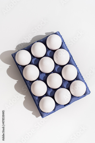 Una docena de huevos blancos de gallina en una huevera de cartón azul sobre una mesa blanca. Vista superior photo