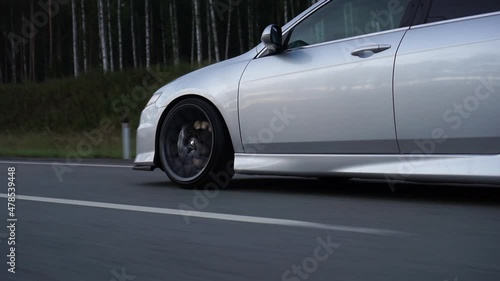Side view of silver car rides on summer highway. Footage. Perfect auto drives fast on country road. Big sports alloy wheels with powerful brakes