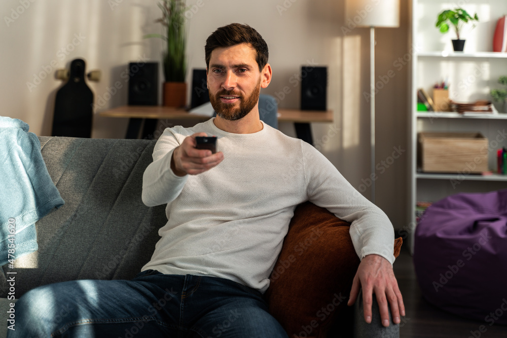 Man dressed in casual clothes lying on the sofa in home and holding remote control while watching TV Man dressed in casual clothes lying on the sofa in home and holding remote control while watching TV