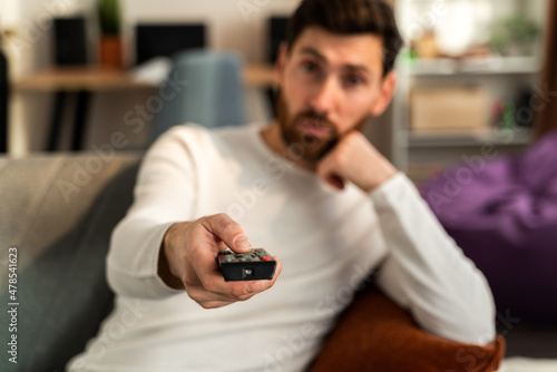 Bored young caucasian man holding tv remote control while sitting on a couch at home