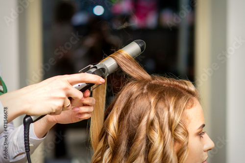 Hairdresser makes curls with a curling iron for the young woman with long brown hair in a beauty salon