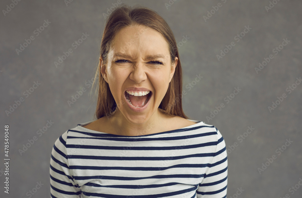 Obraz premium Close up studio shot of a crazy stressed beautiful young woman with an angry displeased face expression screaming loudly at the camera. Portrait of a mad girlfriend or enraged school teacher
