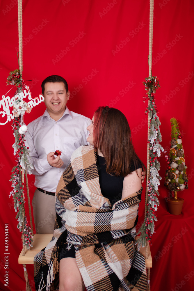 Obraz premium happy couple hugging on wooden swing decorated with christmas tree branches on red background. cozy young family.