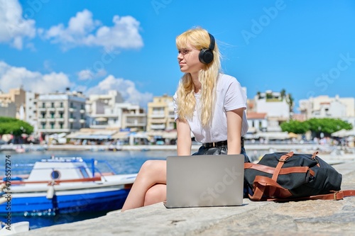 Female teenager in headphones with laptop sitting near the water, sea harbor with boats