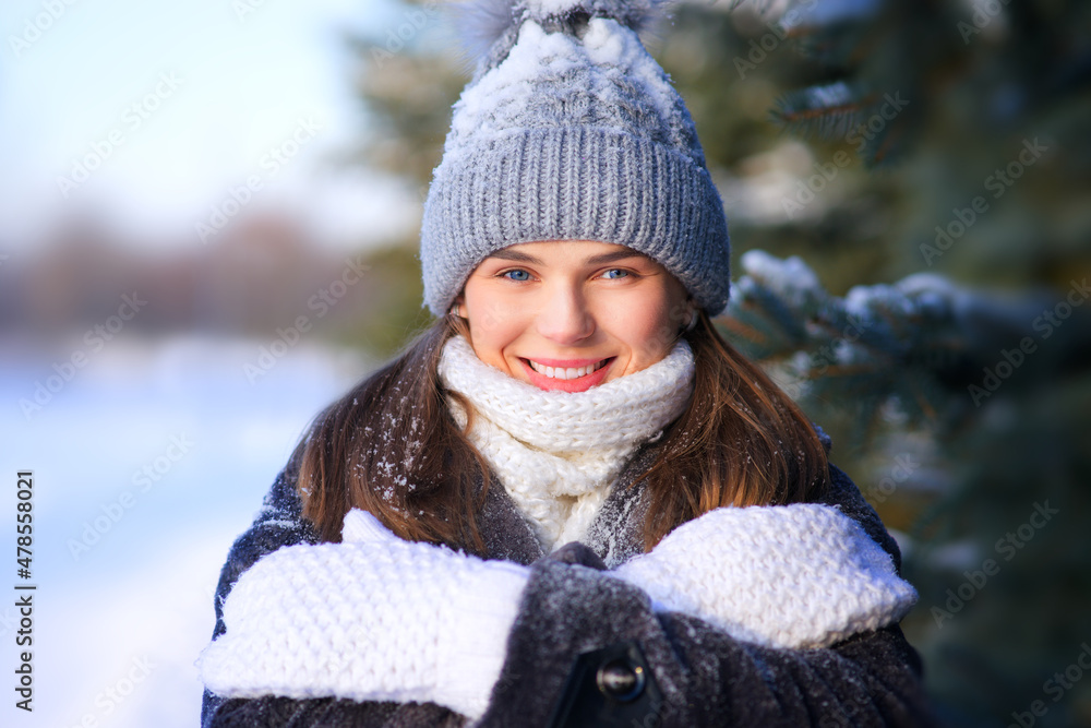 Obraz premium Portrait of happy positive girl, young beautiful attractive pretty woman is freezing walking outdoors in winter snowy park at cold frosty day in hat, scarf, gloves in snow, smiling, looking at camera