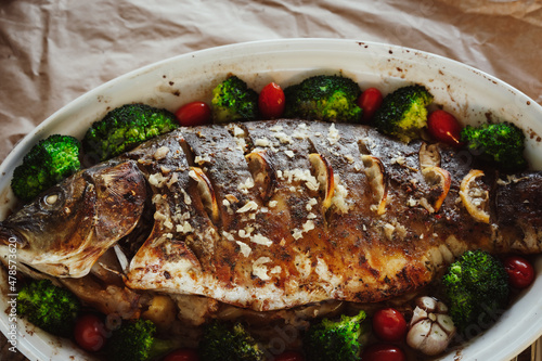 Baked fish carp with apples, broccoli, cherry tomatoes, garlic, lemon and herb spices in baking pan. Healthy eating concept.