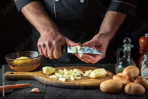 Before making the fries, the chef uses a knife to cut the raw potatoes into small pieces. Close-up of a cook hands while working in a restaurant kitchen