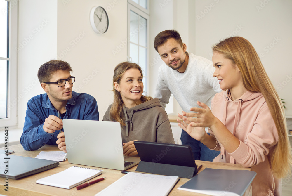 © Studio Romantic - Group of happy smart university students sitting at desk in classroom, working on project together, using laptop and tablet, discussing business startup ideas, doing Internet research on the subject