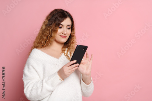 A cheerful woman, standing isolated on a pink background with a smartphone in her hands.