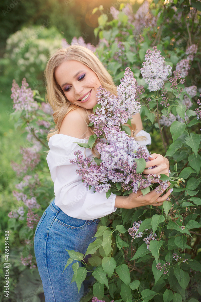 a woman with blond hair in a white blouse and jeans hugs a lilac bush a woman with blond hair in a white blouse and jeans hugs a lilac bush