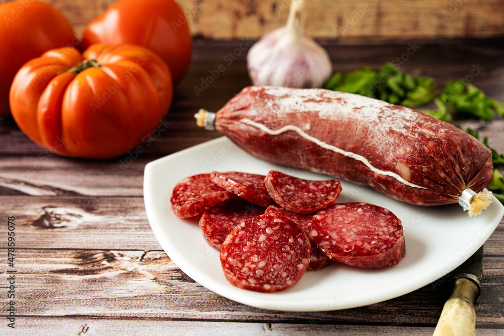 View of whole salami chopped and cut into thin slices on a white plate on a rustic wooden table. Rustic country food concept.