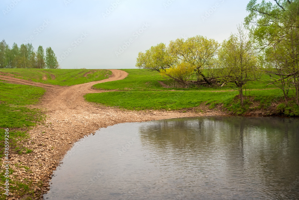 Rural dirt road in rural areas. Green fields, trees and a small river. Beautiful spring landscape.