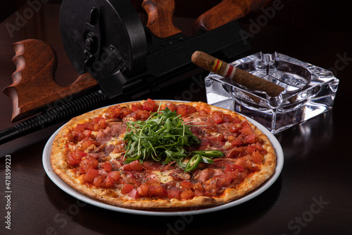 Thompson submachine gun on a dark brown table and ham pizza and a Cuban cigar