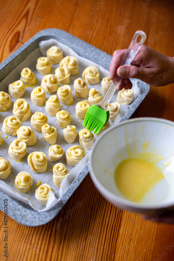 Smearing the dough with egg yolk and silicone brush. There is an egg yolk in the plate. Baking in the shape of a rose or snail. Home cooking. Close-up. Wooden table. Smearing the dough with egg yolk and silicone brush. There is an egg yolk in the plate. Baking in the shape of a rose or snail. Home cooking. Close-up. Wooden table.