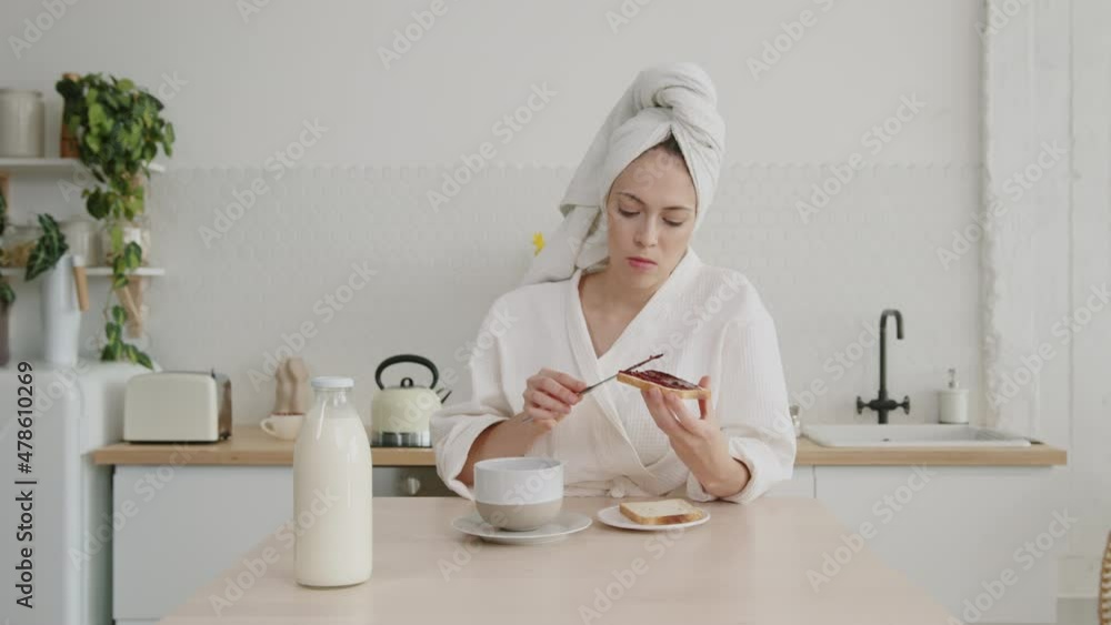 Young woman in the morning at the kitchen table during breakfast in a white robe and with a towel on her head spreads delicious jam on toast and drinks coffee. Young woman in the morning at the kitchen table during breakfast in a white robe and with a towel on her head spreads delicious jam on toast and drinks coffee.