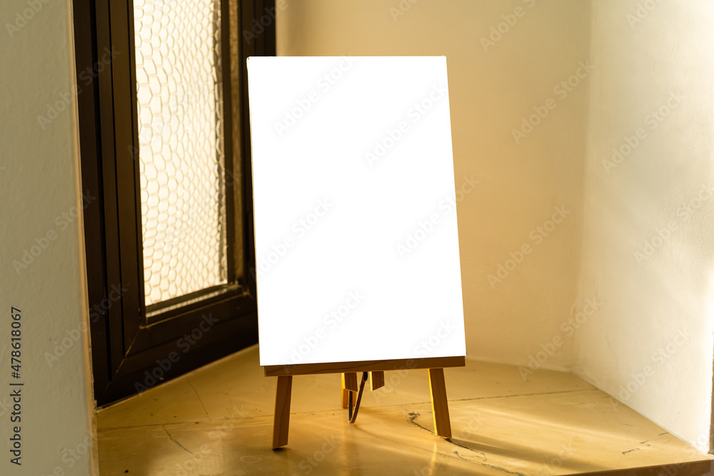wooden easel with white blank canvas stands near the window in an art studio, prepared for artist work. home activities wooden easel with white blank canvas stands near the window in an art studio, prepared for artist work. home activities