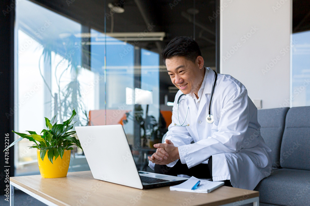 Friendly Asian doctor uses laptop to remotely communicate with patient, man in clinic consults online, looks at monitor screen and webcam Friendly Asian doctor uses laptop to remotely communicate with patient, man in clinic consults online, looks at monitor screen and webcam