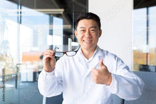 Happy asian ophthalmologist doctor holding glasses, recommending to improve eyesight, looking at camera and holding thumb up approvingly