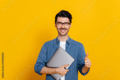 Attractive smart caucasian guy, with glasses, student or freelancer, holding a laptop in hand, standing against isolated orange background, looking at camera, smiling friendly, shows thumb up gesture