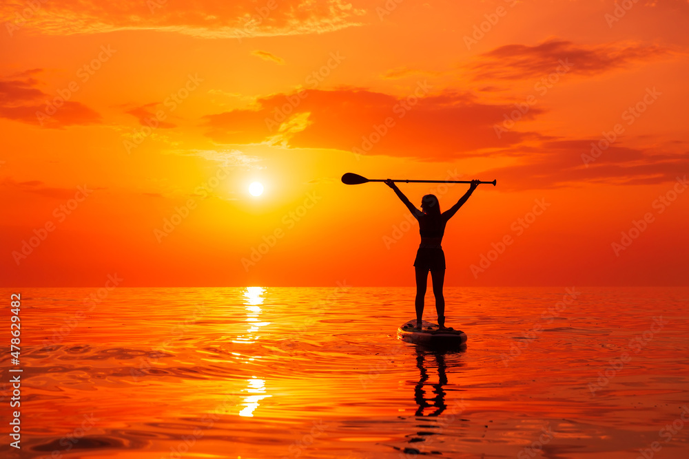 Silhouette of woman on stand up paddle board at quiet sea with sunset or sunrise. Woman posing on SUP board and bright sunset