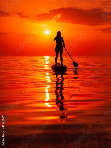 Silhouette of woman on stand up paddle board at quiet sea. Woman posing on SUP board and bright sunset with reflection