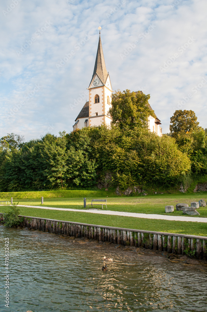 Sunlit church in the village of Maria Worth on the shores of Lake Worthersee in the Austrian mountains. High church clock tower, park with benches and waterfront. Sunlit church in the village of Maria Worth on the shores of Lake Worthersee in the Austrian mountains. High church clock tower, park with benches and waterfront.