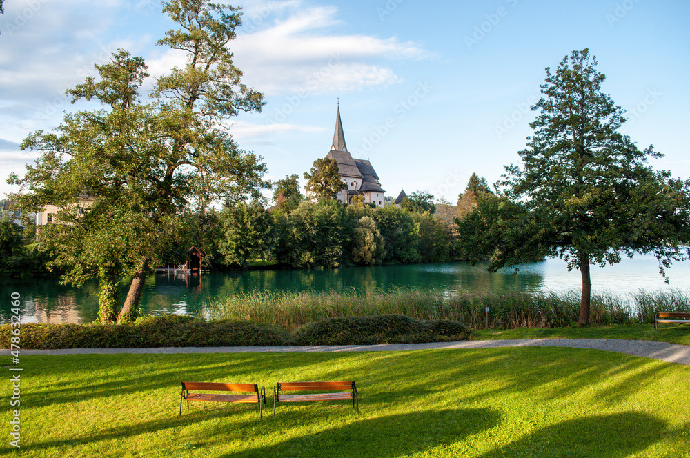 Naklejka premium Sunlit church in the village of Maria Worth on the shores of Lake Worthersee in the Austrian mountains. Irradiated grass in the park and two benches. Beautiful lake with clear blue water.