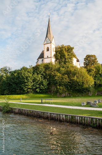 Sunlit church in the village of Maria Worth on the shores of Lake Worthersee in the Austrian mountains. High church clock tower, park with benches and waterfront.