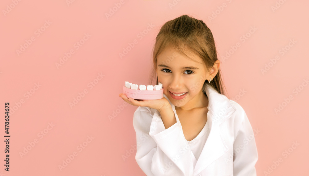 Cute little baby girl demonstrates a healthy snow-white smile, holding a model of a jaw in her hand to demonstrate proper brushing of teeth. International Dentist's Day. A child in a medical gown Cute little baby girl demonstrates a healthy snow-white smile, holding a model of a jaw in her hand to demonstrate proper brushing of teeth. International Dentist's Day. A child in a medical gown
