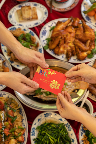 Lunar New Year flat lay of two hands exchanging a red envelope above traditional Chinese food and offering on a table top.
