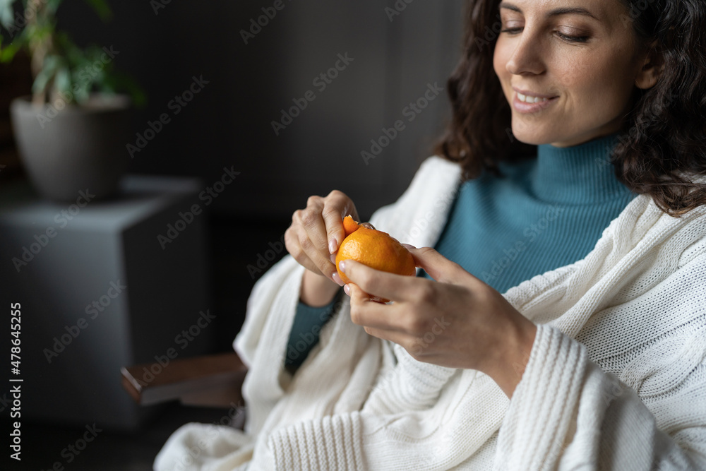 Woman with fresh fragrant tangerine at home. Selective focus of happy cheerful female covered with plaid peeling juicy citrus fruit while relaxing indoors, eating vitamin C rich food in winter season Woman with fresh fragrant tangerine at home. Selective focus of happy cheerful female covered with plaid peeling juicy citrus fruit while relaxing indoors, eating vitamin C rich food in winter season