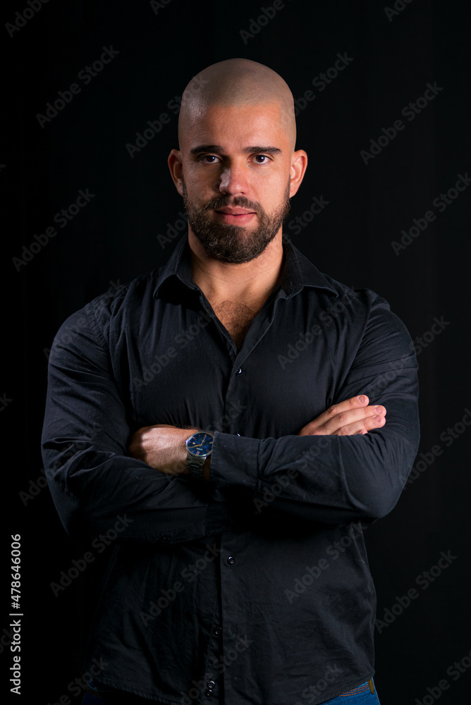  Retrato de homem careca jovem em estúdio com fundo preto. homem vestindo camisa social preta, com braços cruzados, olhando sério para a câmera.