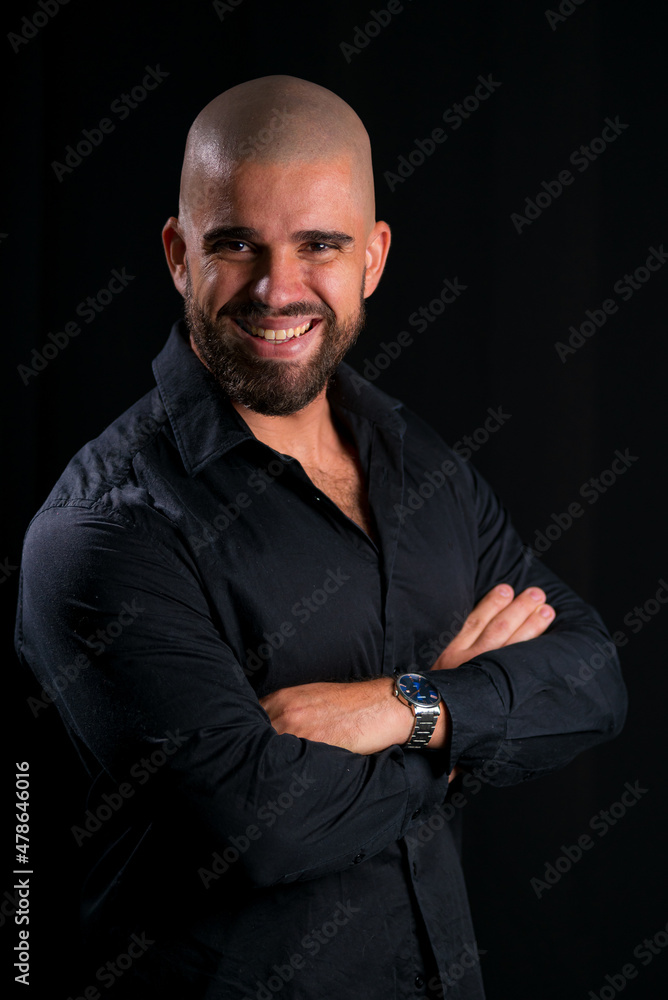 Retrato de homem careca jovem em estúdio com fundo preto. homem vestindo camisa social preta, com braços cruzados, olhando para a câmera e sorrindo.