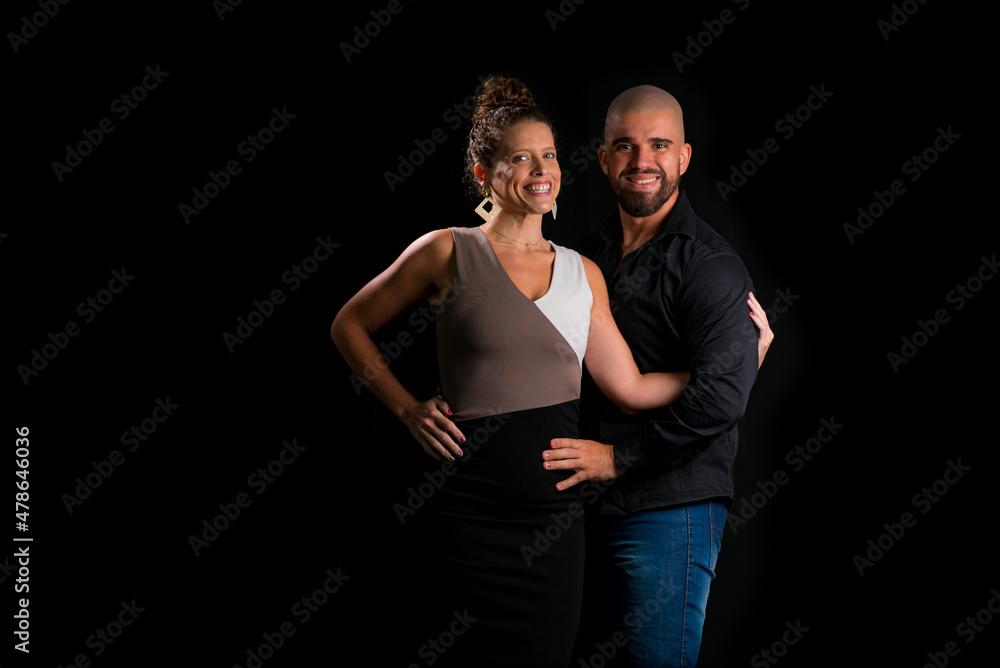 Obraz premium Retrato de casal jovem em estúdio com fundo preto. Mulher loira vestindo vestido social e homem branco careca vestindo camisa social preta e jeans. Abraçados olhando para a câmenta.