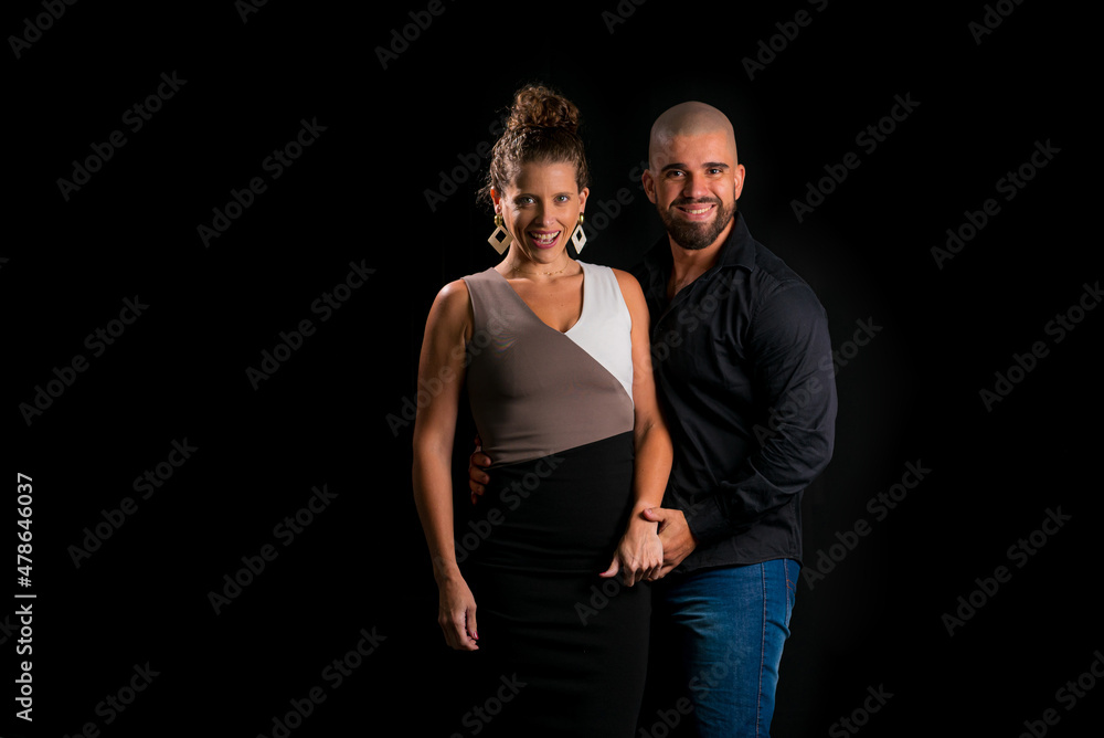Obraz premium Retrato de casal jovem em estúdio com fundo preto. Mulher loira vestindo vestido social e homem branco careca vestindo camisa social preta e jeans. Homem e mulher sorrindo para a câmera.