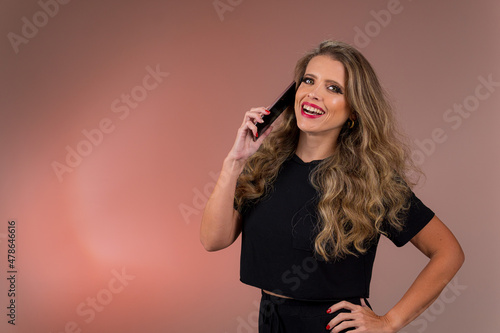 Retrato de mulher loira de olhos verdes, cabelo cacheado, roupa preta em fundo cor salmão no estúdio. Jovem olhando para a câmera falando ao telefone e sorrindo. photo