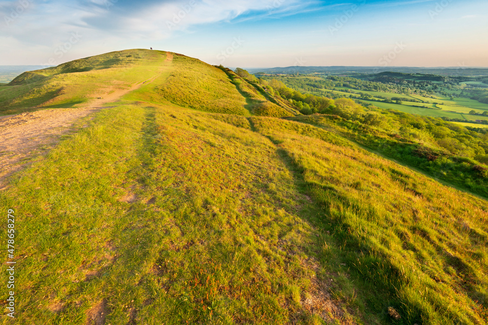 Obraz premium The summer sun rising over Worcestershire countryside,shining low angled sunlight across lush grass on the Malvern hillsides,a distant pathway runs across the peaks,surrounded by beautiful scenery.