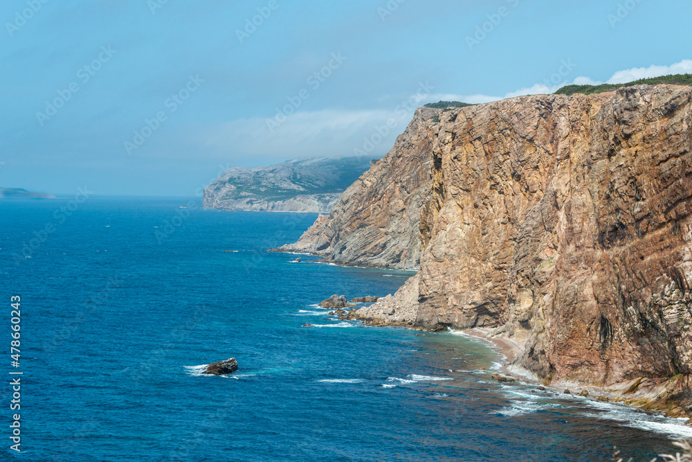 A large rocky mountain of green trees and rocky cliffs. The hill has a ragged and rough coastline. The ocean is blue and smooth below the mountain. The background is blue sky and white fluffy clouds.