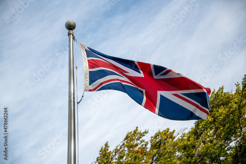 The European flag is known as the Union Jack flying on a metal pole with blue sky and clouds in the background. The red, white, and blue flag is actually three crosses representing the United Kingdom.