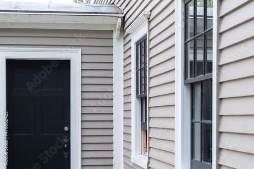 Two vintage double hung windows with a black door on a beige color exterior wooden wall of a house. The windows are dark green with white trim. There's snow on the ground and steps of the entrance.