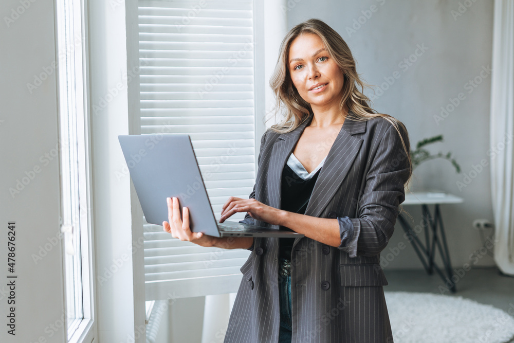 Young smiling blonde woman with long hair in stylish grey suit working at laptop in the bright modern office Young smiling blonde woman with long hair in stylish grey suit working at laptop in the bright modern office